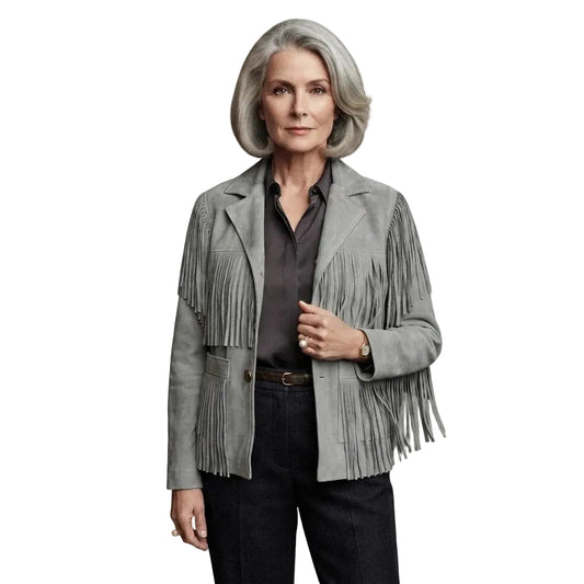 A woman with gray hair wearing a light gray suede fringe jacket over a dark button-down shirt and dark pants, photographed against a plain white background.