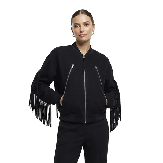 A woman modeling a black bomber jacket with long fringe detailing on the sleeves, paired with black pants, against a plain light gray studio background.
